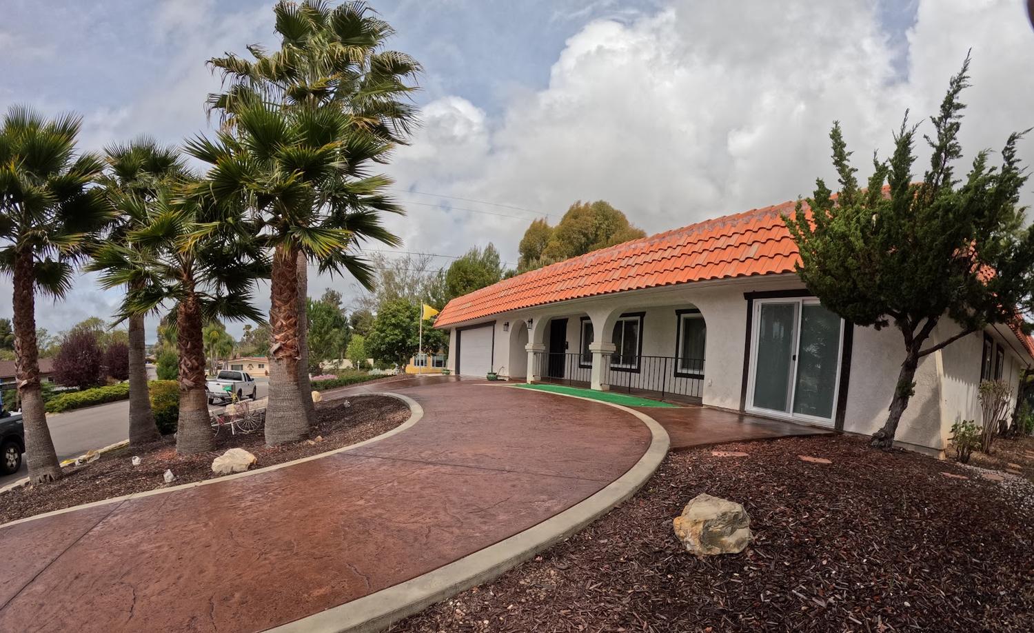 A stamped and stained driveway in warm red-brown curving past a row of palm trees to a house with a red tile roof.