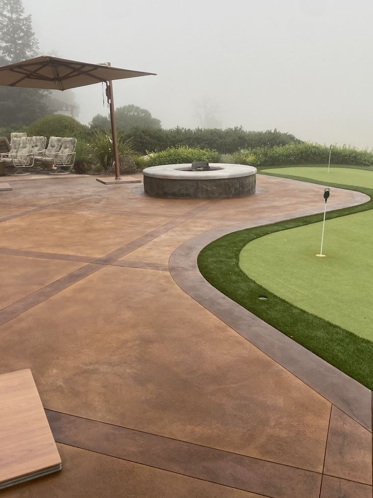 A stained concrete patio curving around a putting green, with a round fire pit at its center.
